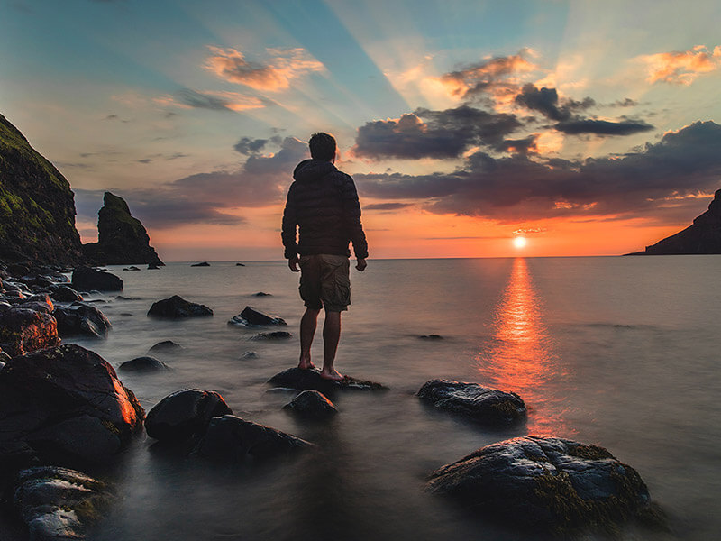 Man standing on rock overlooking the ocean with sunset in the background