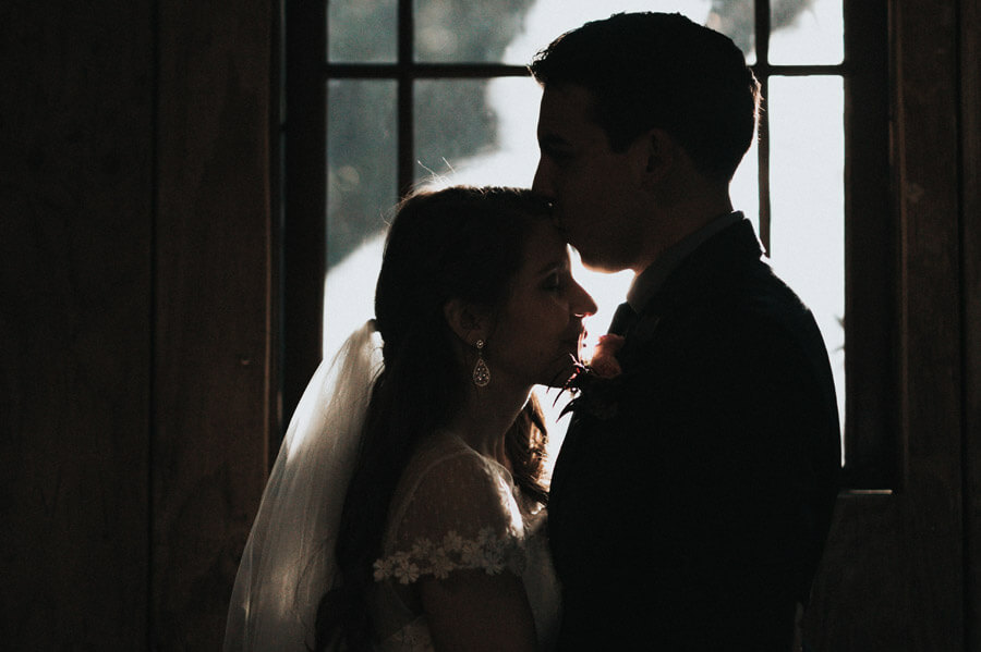 Groom kisses bride on the forehead standing in front of a window